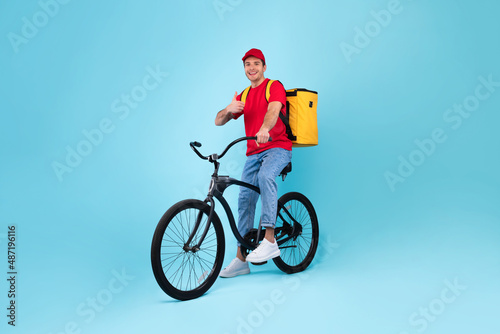 Fototapeta Naklejka Na Ścianę i Meble -  Cheerful Male Courier With Bike Gesturing Thumbs Up, Blue Background