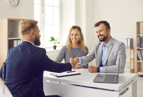 Happy young couple shaking hands with real estate agent after signing contract at his office. Clients smiling and exchanging handshake with realtor. Happy family thanking loan advisor for consultation