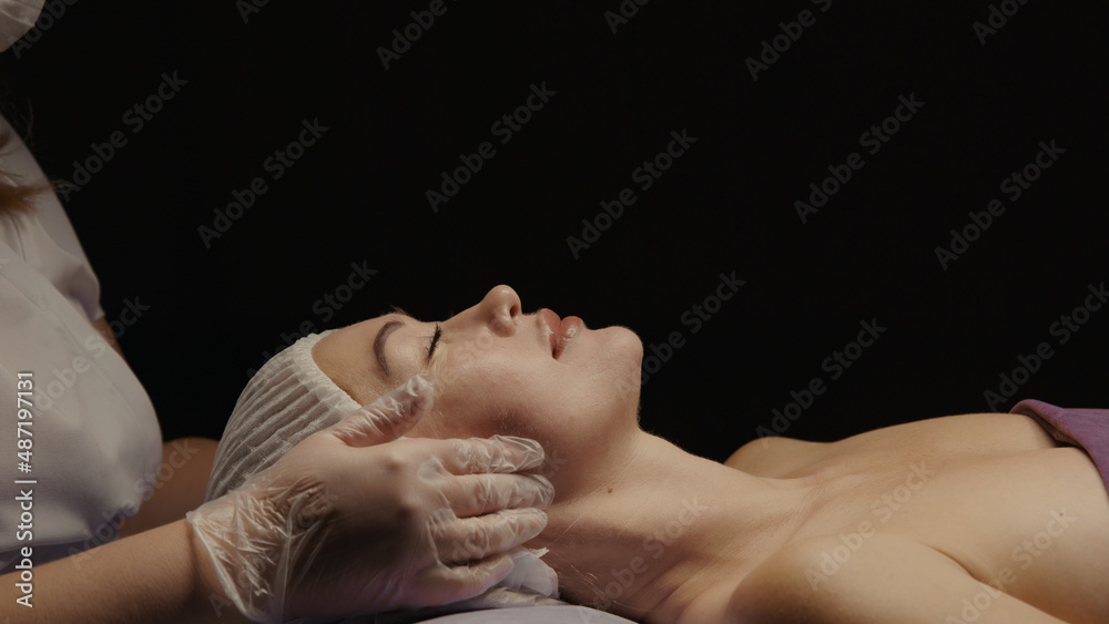 Young hypnotic face woman receiving anti-ageing facial massage in spa salon relax. Wellness body skin care face beauty treatment, rejuvenation procedure getting head face massage. Black background