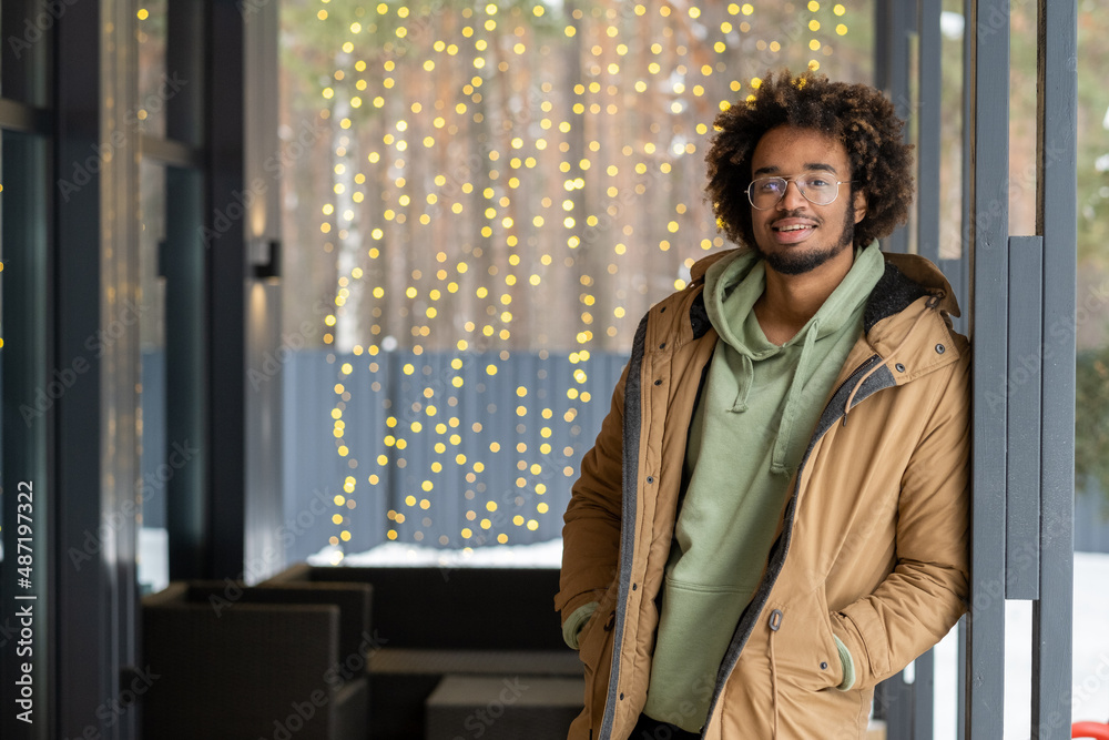 Portrait of African guy in warm coat smiling at camera while standing on the porch of the house in winter day Portrait of African guy in warm coat smiling at camera while standing on the porch of the house in winter day