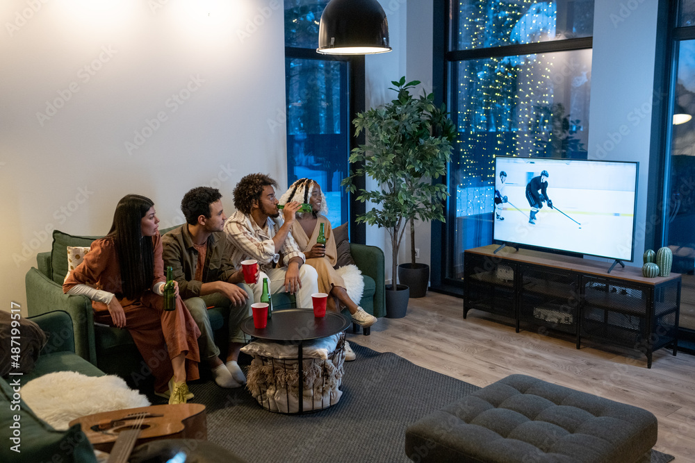 Naklejka premium Young friends sitting on sofa with beer and watching sport match on TV in a big modern apartment