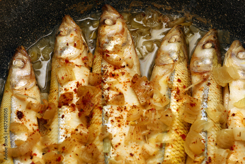 Delicious appetizing fresh fried european smelt fish with white onion and red seasoning on black non-stick frying pan, top view close-up. Six fish lie side by side, cooking at home photo