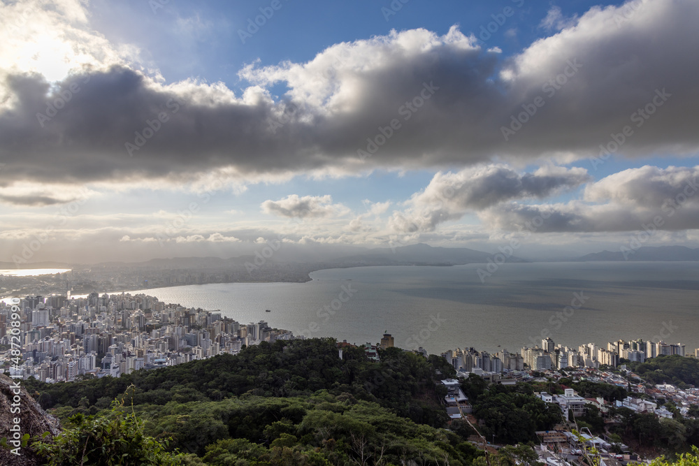 Vista da cidade de Florianópolis e o continente ao fundo com a ponte interligando os lugares. Paisagem com bastante Sol e nuvens.
