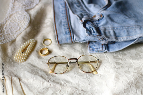 Vintage jeans, white blouse, retro eyeglasses, pearl barrette and gold rings. Fashionable outfit layout. Selective focus.