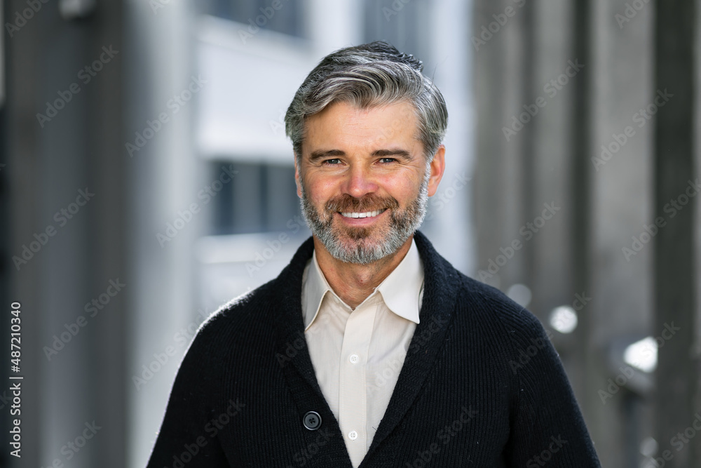 Naklejka premium happy mature man looking at camera on the street. Senior businessman with a beard feels confident. Close-up face of a business man smiling on the background of the building.