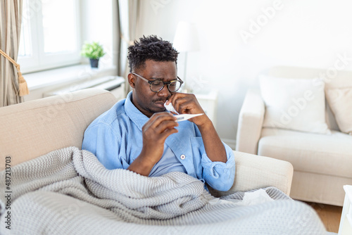 Ill african young man covered with blanket blowing running nose got fever caught cold sneezing in tissue sit on sofa, sick allergic black man having allergy symptoms coughing at home, Corona 19