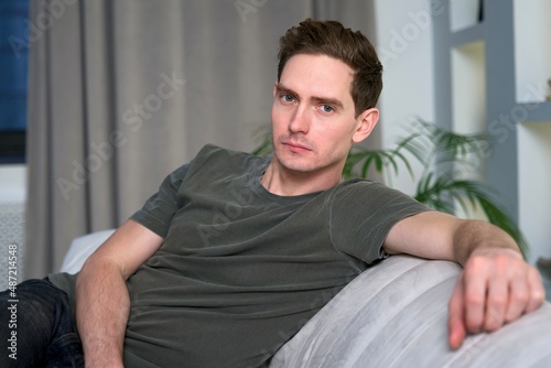 Portrait of serious handsome guy, young relaxed man is sitting on couch or sofa at home or flat in living room looking at camera, resting, relaxing in the evening.