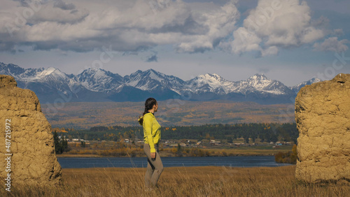 The woman in yellow green sportswear. The traveler near old stone enjoying highland landscape. Tourist are walking against the backdrop of snow-capped mountains. Strong wind.