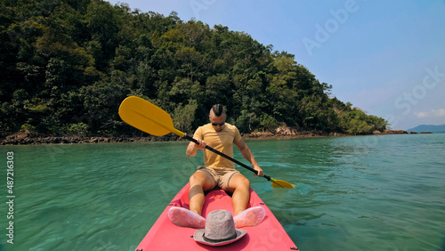 Young man with sunglasses and hat rows pink plastic canoe along sea against green hilly islands with wild jungles. Traveling to tropical countries. Strong guy is sailing on kayak in ocean, front view.