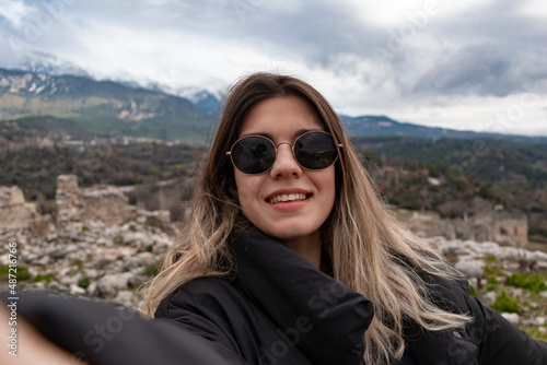 Happy beautiful girl takes a selfie portrait on vacation outdoor. She has sun glasses and winter clothes. Travel alone concept. © burhan