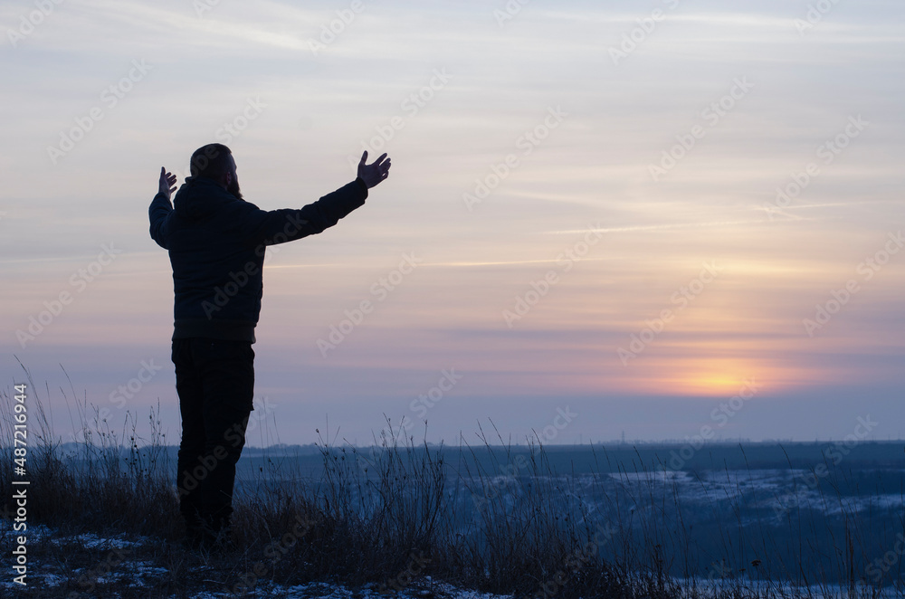 Fototapeta premium Pray. Repentance. Silhouetted men on a background of blue sky and sunset. Kneeling Prayer to God. Glorification. Praising God. A man with his hands up. Worship God 