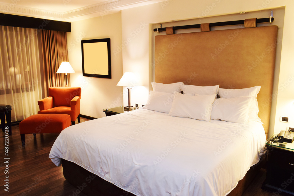 Habitación de hotel, cama, muebles, almohadas, sabanas blancas, sillas y sofá. Vista lateral