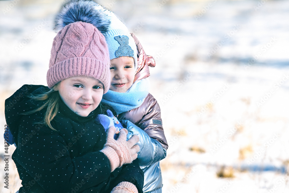 Obraz premium Portrait of two beautiful happy little girls on a walk in a winter park in warm clothes, hats and scarves