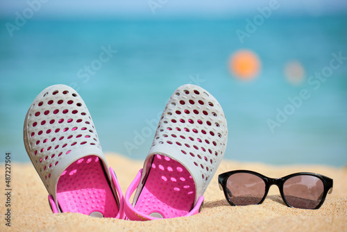 Closeup of clogs shoes and black protective sunglasses on sandy beach at tropical seaside on warm sunny day. Summer vacation concept