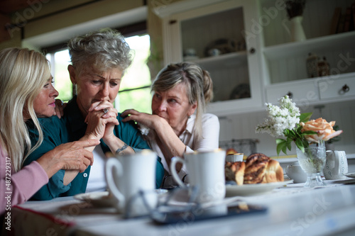 Female senior friends consloing and supporting sad friend indoors at home.