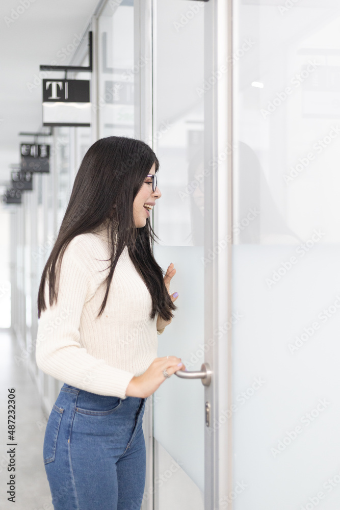 White woman standing on an office corridor smiling and waving through the glass