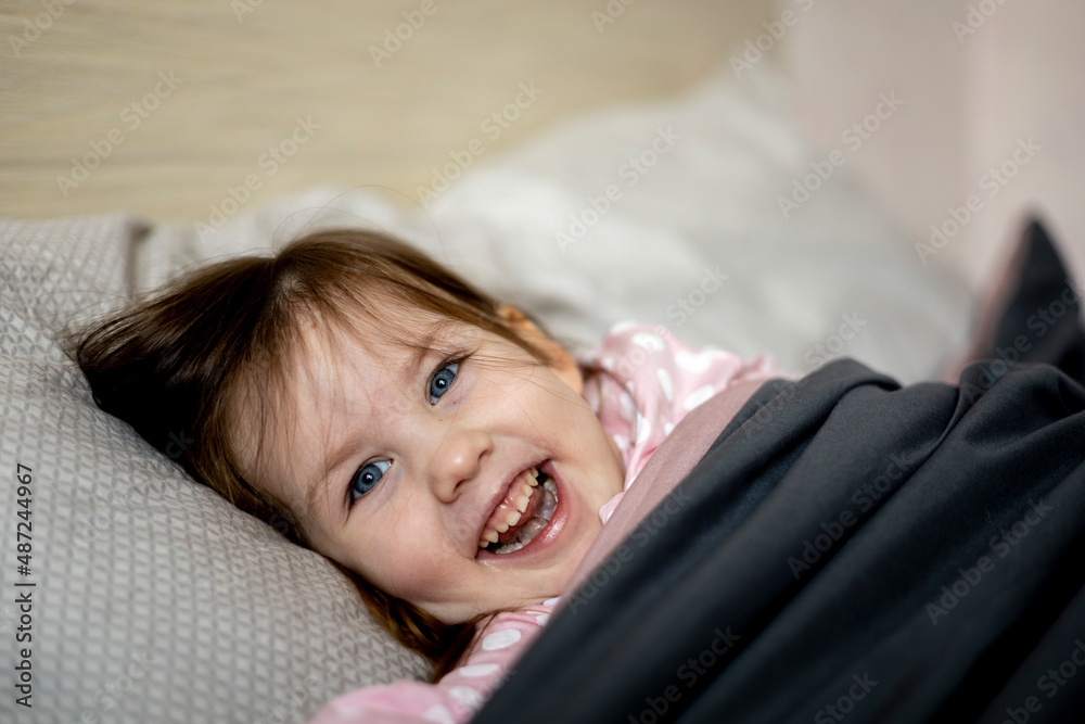 Little dark-haired girl in pink pajamas lies on a cozy soft bed. Good morning. Little dark-haired girl in pink pajamas lies on a cozy soft bed. Good morning.