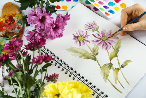 Woman painting chrysanthemums in sketchbook and flowers at white table  closeup