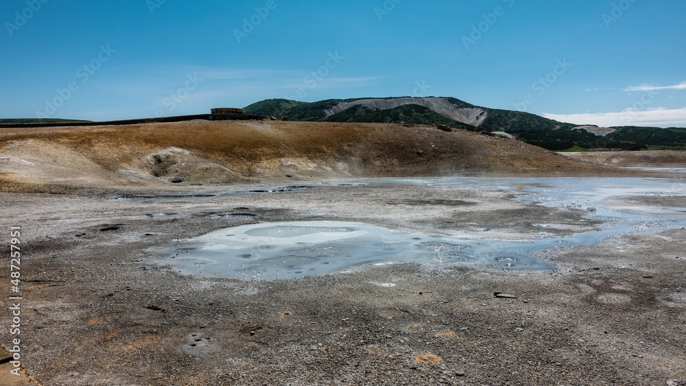The water in the thermal springs is blue and opaque. Sulfur deposits are visible on the surrounding soil. A mountain range against an azure sky. Kamchatka. The caldera of the extinct volcano Uzon The water in the thermal springs is blue and opaque. Sulfur deposits are visible on the surrounding soil. A mountain range against an azure sky. Kamchatka. The caldera of the extinct volcano Uzon