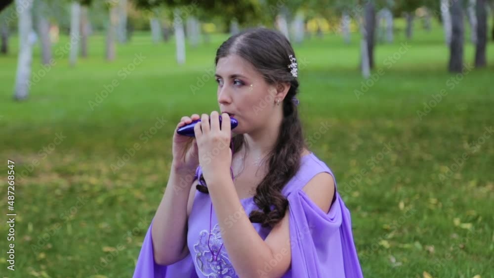 A young attractive woman with make-up in a medieval dress plays the ocarina sitting on the grass against the backdrop of a park, forest. A young attractive woman with make-up in a medieval dress plays the ocarina sitting on the grass against the backdrop of a park, forest.