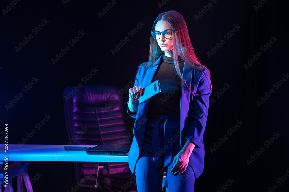 Naklejka premium Woman entrepreneur. Businesswoman in her own office. Businesslady is sitting at office desk. Stylish businesswoman. Girl in blue suit is holding phone. Business lady in dark office. Successful girl