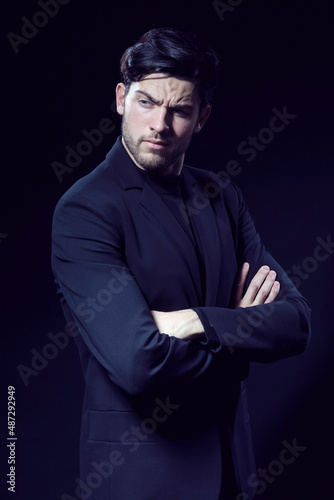 Portrait of Handsome Caucasian Brunet Businessman Wearing Black Suit Posing With Folded Hands Against Black Studio Background.