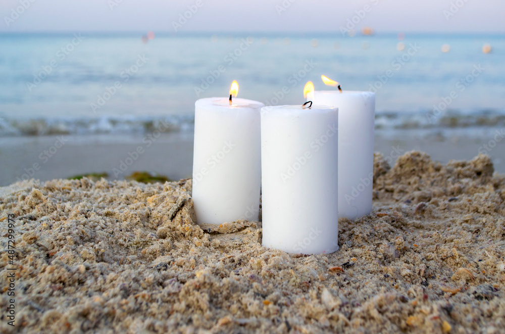 Three large white paraffin candles burn on sand beach background sea waves.  Decorative Burning candles on sea coast close up. Candlelight, candle  flame. Romantic mood backdrop. Concept date relaxation Stock Photo