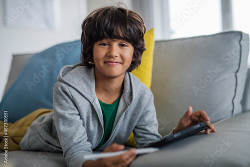 Little multiracial boy with tablet lying on sofa at home, lookingat camera. photo
