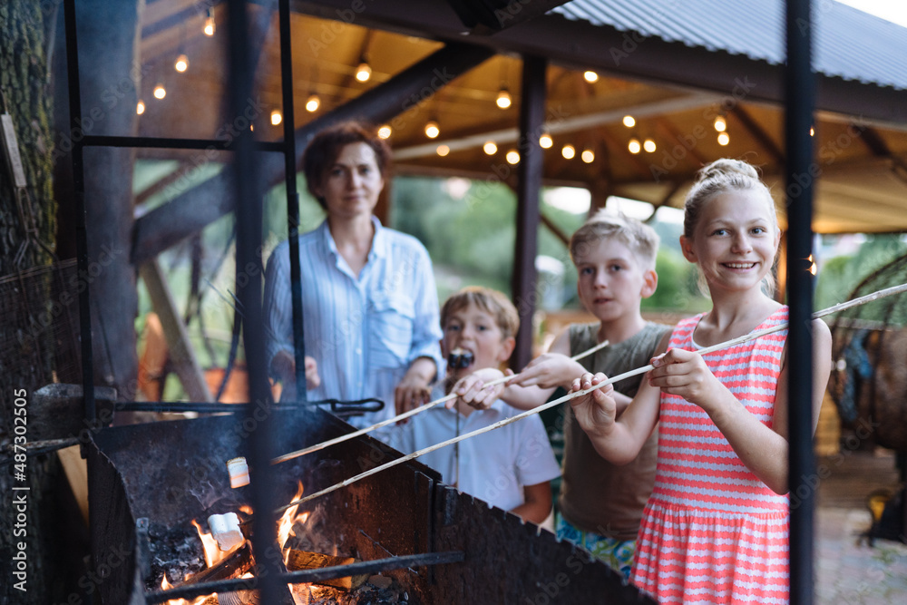 Family with two children cooking marshmallow candies on barbecue on the grill brazier at backyard. Children Roasting Marshmallows at Summer Evening.
