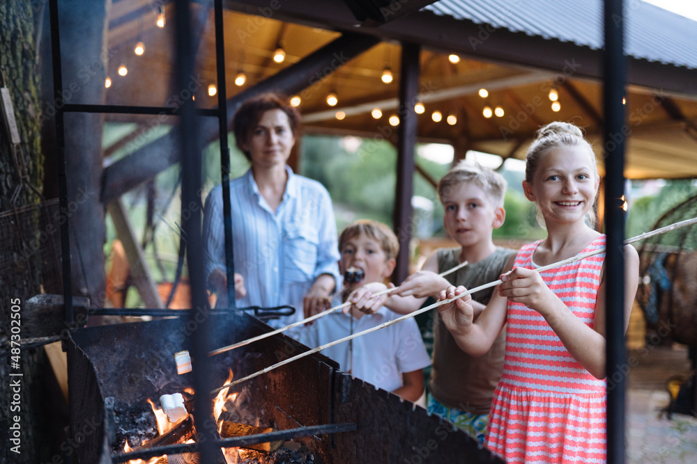 Family with two children cooking marshmallow candies on barbecue on the grill brazier at backyard. Children Roasting Marshmallows at Summer Evening. Family with two children cooking marshmallow candies on barbecue on the grill brazier at backyard. Children Roasting Marshmallows at Summer Evening.