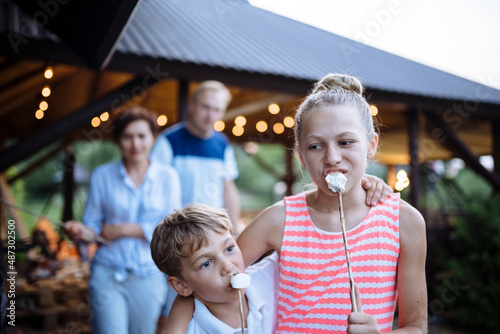 Family with two children cooking marshmallow candies on barbecue on the grill brazier at backyard. Portrait of boy eating roasts marshmallow on stick.