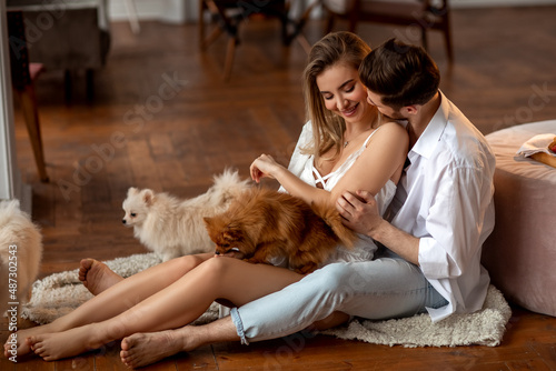 Young caucasian affectionate couple at home in white clothes spend weekend together  play with their puppies. Positive  joy  happiness  love concept