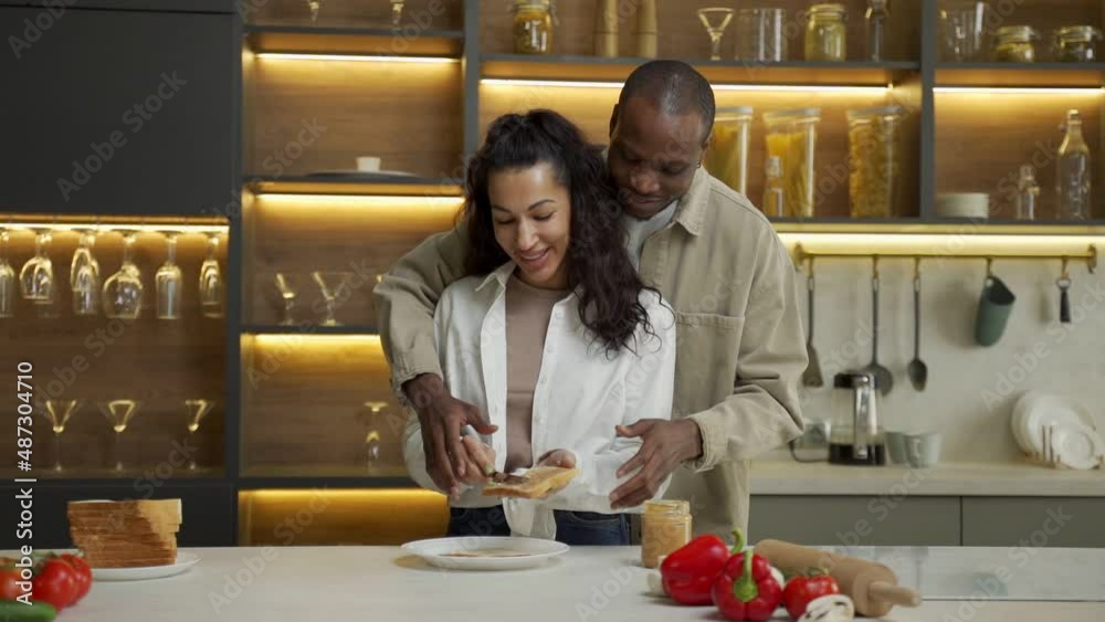 African American man helps beloved mixed race woman spread sauce on bread in modern kitchen. Happy couple hugs cooking dinner against countertop slow motion