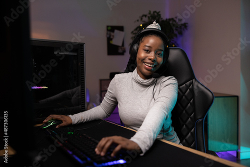 Smiling young woman with dark skin hair tied up in ponytail headset playing professionally on computer online games with team members gaming accessories neon led backlit keyboard mouse room at night © ABCreative