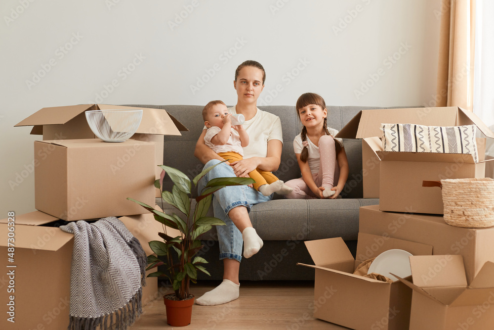 Indoor shot of young woman wearing casual style attire sitting on floor with her children, looking at camera, relocating to a new apartment, surrounded with cardboard boxes. Indoor shot of young woman wearing casual style attire sitting on floor with her children, looking at camera, relocating to a new apartment, surrounded with cardboard boxes.