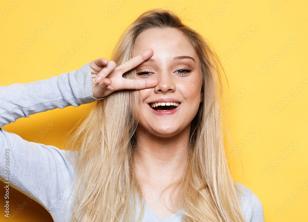 happy blond woman showing victory sign and looking at camera isolated over yellow background happy blond woman showing victory sign and looking at camera isolated over yellow background