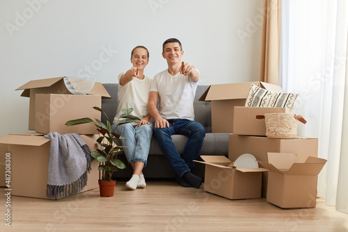 Indoor shot of excited happy young couple sitting on sofa surrounded with carton boxes with personal belongings, pointing to camera, choosing you for buying new apartment, mortgage. © sementsova321
