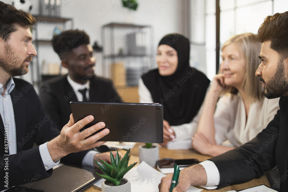 Multi ethnic team of business partners looking on one tablet screen during brainstorming at conference room. Five people discussing common working project at office. Multi ethnic team of business partners looking on one tablet screen during brainstorming at conference room. Five people discussing common working project at office.