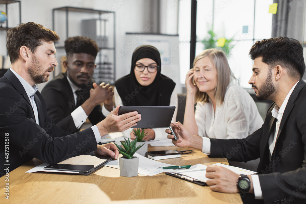 Multi ethnic group of business people wearing stylish formal wear using digital tablet during briefing at boardroom. Modern technology and cooperation concept. Multi ethnic group of business people wearing stylish formal wear using digital tablet during briefing at boardroom. Modern technology and cooperation concept.