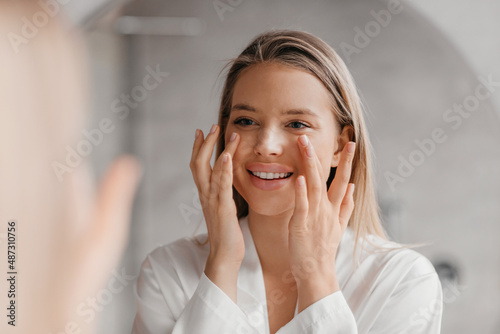 Pretty lady making face-lifting massage in bathroom, looking at mirror and smiling, using moisturizing face cream photo