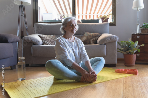 Elderly caucasian woman doing yoga exercises and stretching on a mat in the living room of her home