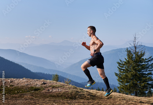 Side view of professional trail runner who taking part in summer marathon of running on mountain hills. Man with naked athletic body running against the backdrop of mountain panorama.