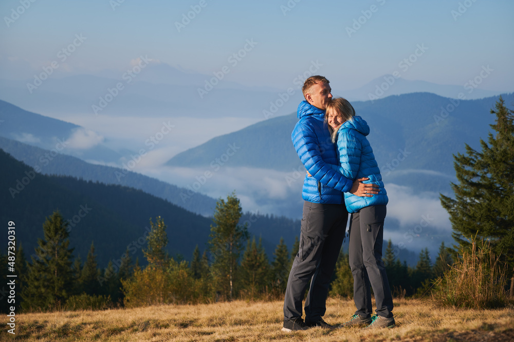 Happy couple hugging and smiling while standing on grassy hill with blue sky and mountains on background. Young man traveler embracing his charming wife while enjoying beauty of nature in mountains.