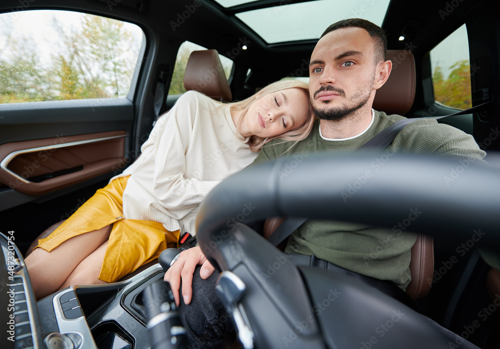 Happy man and woman traveling in car. Beautiful young couple sitting on the front passenger seats, handsome man driving comfortable automobile. Tired woman laying on guy's shoulder with closed eyes