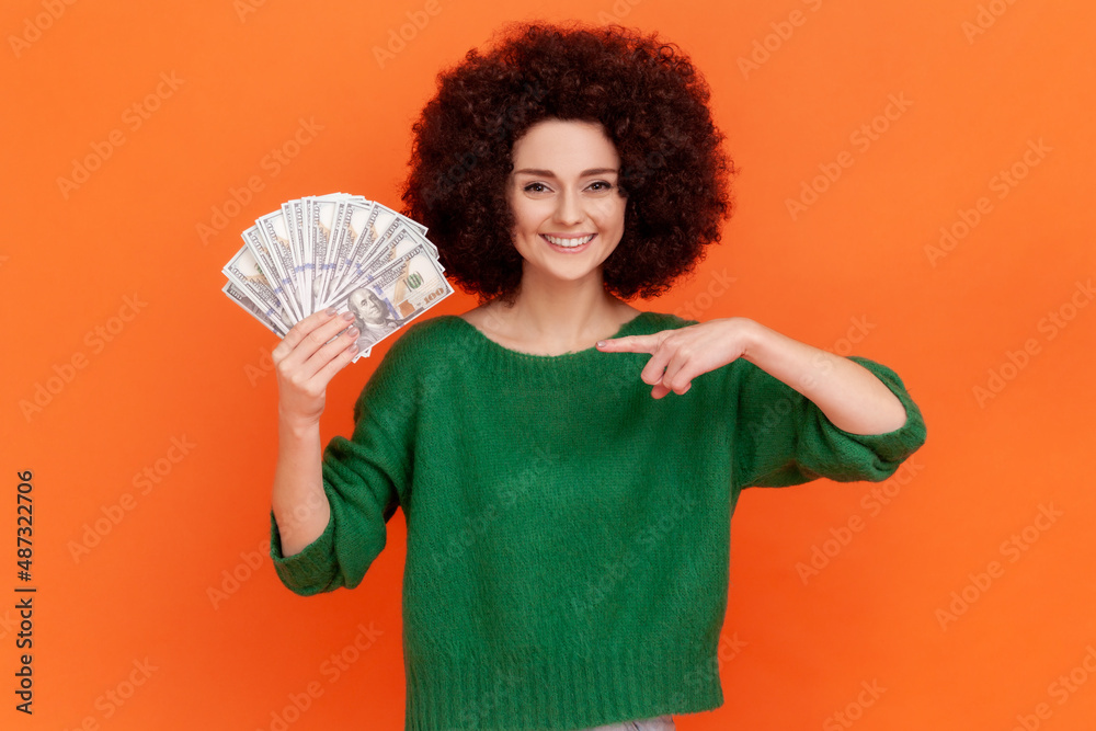 Portrait of gratified rich woman with Afro hairstyle wearing green sweater pointing finger on earned dollar banknotes, enjoying success and big profit. Indoor studio shot isolated on orange background