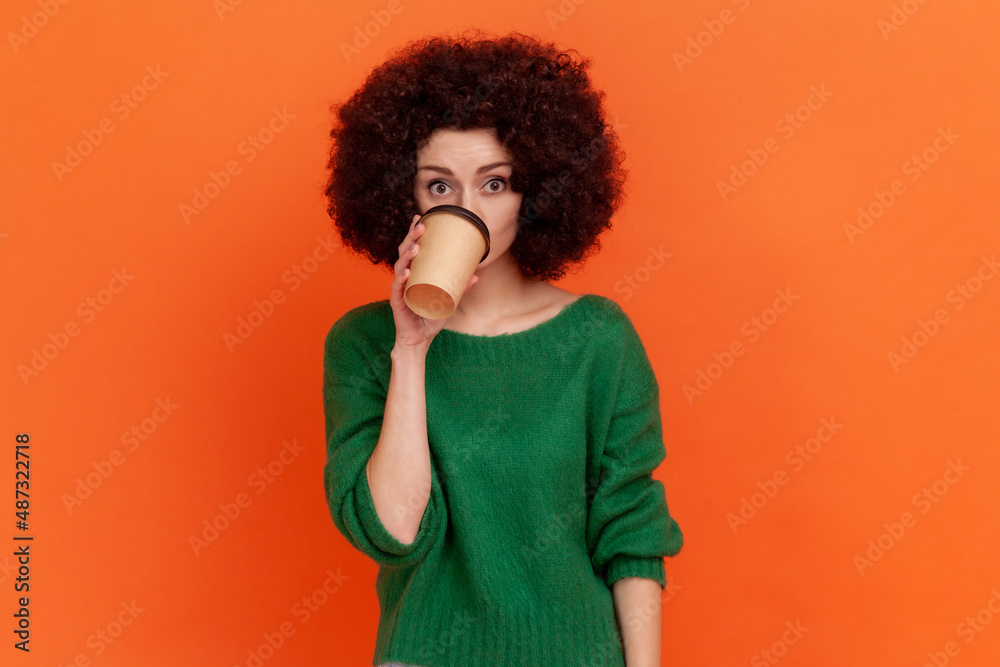 Good looking woman with Afro hairstyle in green casual sweater standing drinking hot beverage, take away coffee from disposable cup, needs energy. Indoor studio shot isolated on orange background. Good looking woman with Afro hairstyle in green casual sweater standing drinking hot beverage, take away coffee from disposable cup, needs energy. Indoor studio shot isolated on orange background.