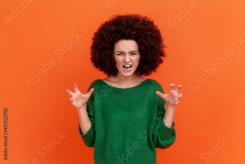 Portrait of angry woman with Afro hairstyle wearing green casual style sweater expressing aggressive emotions, raised arms, screaming. Indoor studio shot isolated on orange background.