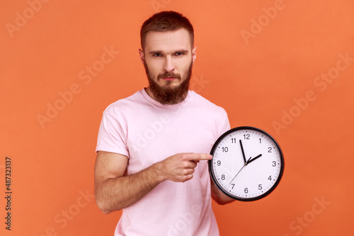 Portrait of serious bearded man pointing finger at big wall clock looking at camera, worried of completed on time work, wearing pink T-shirt. Indoor studio shot isolated on orange background.