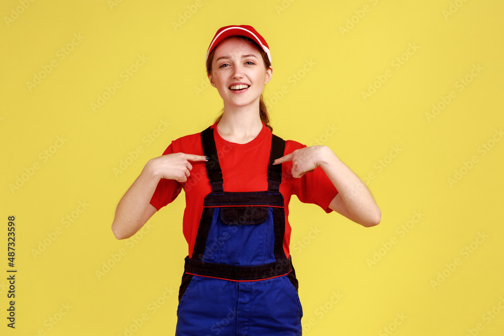 Portrait of happy delighted worker woman standing and pointing at herself, being satisfied of result of her work, wearing overalls and red cap. Indoor studio shot isolated on yellow background. Portrait of happy delighted worker woman standing and pointing at herself, being satisfied of result of her work, wearing overalls and red cap. Indoor studio shot isolated on yellow background.