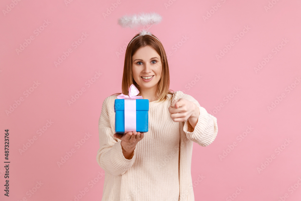 Portrait of winsome young blond woman with halo over head holding blue gift box in hands and pointing at camera, wearing white sweater. Indoor studio shot isolated on pink background. Portrait of winsome young blond woman with halo over head holding blue gift box in hands and pointing at camera, wearing white sweater. Indoor studio shot isolated on pink background.